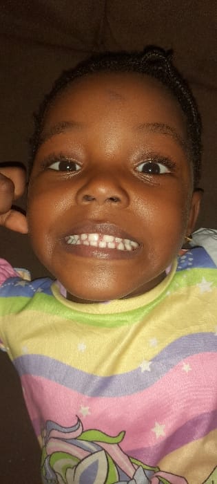 This image captures a close-up, frontal view of a young African child, likely between two and five years old, located indoors within the city of Ashipa, Nigeria. The child exhibits dark skin, dark eyes, and dark hair styled in short braids across the forehead. The child is looking directly at the camera with a subtle smile, exposing upper and lower primary teeth, notably featuring a central diastema between the upper incisors. A dark-skinned hand is partially visible on the left side of the child's face, near the ear. The child is dressed in a multi-colored garment with horizontal stripes in pastel shades of light green, yellow, purple, and pink, embellished with scattered white star patterns. A portion of a graphic depicting a stylized mythical creature, possibly a unicorn, in similar pastel tones, is visible on the lower section of the clothing. The background is an undifferentiated dark brown, textured surface, indicative of upholstered furniture. Illumination is consistent, highlighting the child's facial features.
