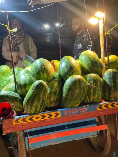 A nighttime outdoor market scene in Kabul, Afghanistan, features a stall laden with a large quantity of watermelons. Two men are visible. The man on the left, wearing a tan garment and a checkered scarf, is partially obscured. The man on the right, wearing a black and white patterned shirt, is also partially visible, looking away from the camera. The watermelons are displayed on a wooden cart with colorful painted details, including a yellow stripe with black chevrons and a partial phone number "0788 718". Overhead lighting, possibly from lamps, illuminates the scene.