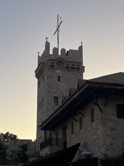 The image shows a stone structure that appears to be a castle or fortified building. The building features crenellated battlements and multiple windows, some of which are dark and appear to be set deep within the stone walls. A wooden roof with visible rafters overhangs a section of the building, and there are wooden balconies or walkways attached to the exterior. A flagpole with crossed arms stands near the top of one of the turrets, with some thin wires extending from it. Small crosses are visible atop some of the turrets. The building is constructed from rough-hewn stone blocks, giving it a rustic and ancient appearance. To the right of the main structure, some greenery, likely trees or bushes, is visible, along with another stone building. The sky is a pale, hazy color, suggesting either early morning or late afternoon light. The overall impression is of a historical site, possibly a medieval castle, set against a muted sky. The image is oriented vertically. No people are visible.