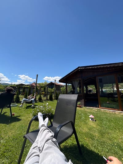 The image shows an outdoor scene, possibly a garden in Polena, Bulgaria, on a sunny day. A person is sitting in a black chair, with legs extended and wearing grey sweatpants and white Adidas socks and sneakers. A wooden structure with glass windows is visible in the background, along with green grass and trees. Another person is sitting further away on a black chair. A woman is seated on the ground wearing jeans and a white shirt with arms raised. Flowers are scattered on the ground. A person is smoking in the upper right.