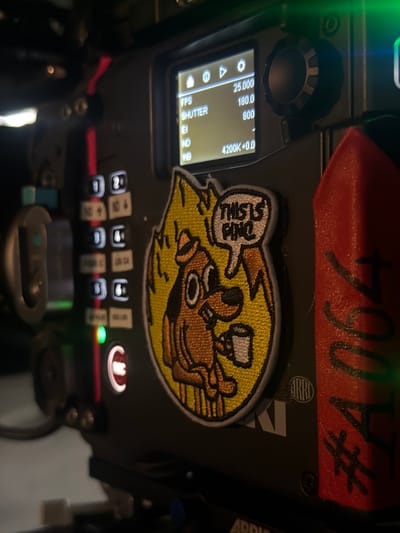 A close-up, low-angle shot captures the top surface of professional film equipment, likely a camera or a monitoring unit, in an indoor setting. The device is predominantly black, featuring a digital display, various controls, and personal adornments.
On the left, a rectangular screen displays key camera settings: FPS at "25.000", SHUTTER at "180.0", EI at "800", and a white balance of "4200K+0.0". Icons for settings and playback are also visible. Above the screen, there's a textured, dark gray rotary control knob.
To the right and below the screen, an array of illuminated buttons with various symbols and numbers (e.g., '1' through '12') are arranged, suggesting customizable functions. A prominent red button with a white symbol (resembling an 'M') and a glowing green indicator light are also visible.
A notable feature is a yellow embroidered patch affixed to the surface, depicting a cartoon dog-like character sitting calmly amidst flames, with a speech bubble exclaiming "THIS IS FINE". Above this patch, a piece of red tape bears the handwritten inscription "#4064".
Partial brand names are visible, with "ARRI" discernible on the right side of the equipment, indicating a high-end camera manufacturer. Another partial text "ADDI" and a circular logo (possibly "KB" or "HB") are also present. The scene is artificially lit, with a green light casting a glow from the upper left. No specific visual cues confirm the given location of San Sebastián De Los Reyes, Spain, from the image itself.