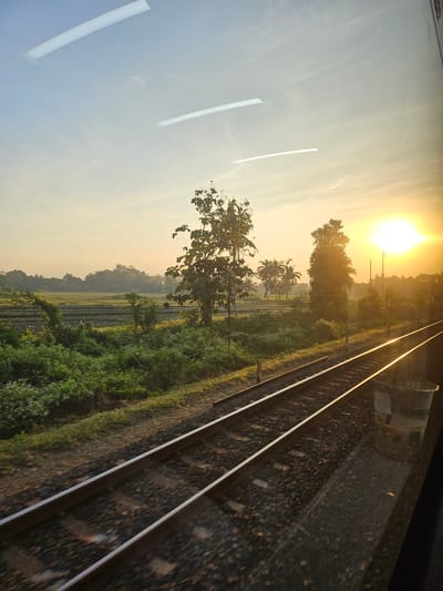 This image depicts a view from a train traveling through Sedayu, Indonesia, during sunrise. The foreground features a set of train tracks, with the right rail glinting under the rising sun. To the left of the tracks, dense green vegetation grows, interspersed with trees and a cultivated field. In the distance, a line of trees forms the horizon, with hills visible beyond them. The sun, a bright, hazy orb, is positioned on the right side of the frame, casting a warm glow over the entire scene. The sky is a pale blue, transitioning to a soft orange near the horizon, with a few wispy clouds. Reflections of fluorescent lights are visible on the window through which the photograph was taken, suggesting an interior view of the train.