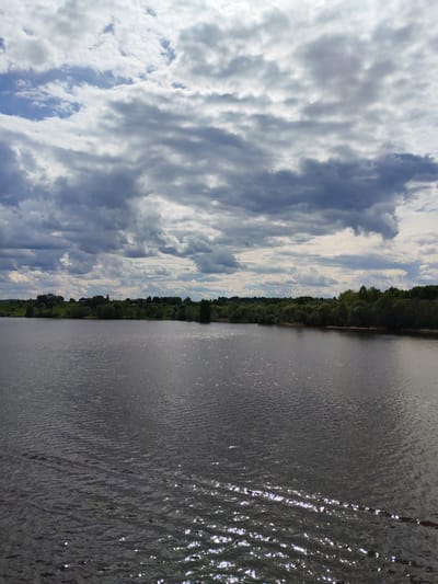 The image depicts a landscape with a body of water, possibly a lake or a wide river, taking up the majority of the frame. The water is dark with surface ripples and reflections of light, creating a sparkling effect. In the background, there is a green shoreline with trees and some visible structures that appear to be buildings, suggesting a settlement. Above the scene, the sky is filled with a mix of light and dark, heavy clouds, indicating potentially unsettled weather. The scene is in the location of Popovo, Russia.