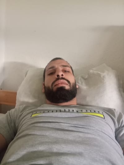 A person with dark hair and a full, dark beard is lying on a bed with a white pillow. They are wearing a gray t-shirt with a yellow stripe and black letters. The background is a plain white wall. The image does not provide any information about the location or city. The location in the city of Domsühl, Germany cannot be verified from the image.
