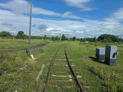 The image shows a set of train tracks in Khurba, Russia, stretching into the distance and surrounded by a grassy field. A wooden utility pole stands to the left of the tracks. Two grey rectangular electrical boxes, each with a red stripe, are positioned on the right side of the frame, adjacent to the tracks. The sky is blue with scattered white clouds. In the background, trees and some distant structures are visible. The scene appears to be a rural or semi-rural area within Khurba.
