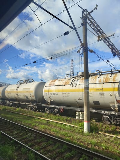 The image shows two adjacent tank cars on a railway track, likely in the city of Фруктовая, Russia, given the text on the cars. The tank cars are white with yellow stripes and appear to be carrying ammonia, indicated by the Russian word "АММИАК" and warning symbols for hazardous materials. The car on the right has the number "782 27816" visible, and other markings such as "20" and "120 KM/H". The car on the left has the number "77957693" and similar markings. Both cars show signs of rust and weathering. In the background, a utility pole with overhead electrical wires for the train system is prominently featured, along with a blue sky with scattered clouds. The foreground shows a grassy area alongside the railway tracks, with more tracks visible. The perspective suggests the photo was taken from inside a moving train, looking out a window.