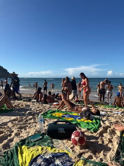 This image depicts a sunny, crowded beach scene in Cairu, Brazil. The foreground features light-colored sand with several beach towels, some displaying the Brazilian flag design in green, yellow, blue, and white. A black JBL portable speaker, a clear plastic water bottle, a small red and white soccer ball, and an open book or tablet are visible on the sand. Numerous individuals are present on the beach and in the shallow ocean waters.

In the mid-ground, a dark-haired woman in a light pink bikini lies prone on a Brazilian flag towel, facing away from the camera. Another woman in a red bikini walks past her, heading towards the water. Other individuals are seated or standing on the sand, sunbathing or conversing. A man in a straw hat and dark shirt walks left across the frame. In the background, many people are wading, swimming, or standing in the calm blue ocean. Small waves are visible near the shore. The sky is clear blue with a few scattered white clouds near the horizon. A green, vegetated hillside or large dune is visible on the far left of the frame. The overall atmosphere suggests leisure and recreation on a clear day.