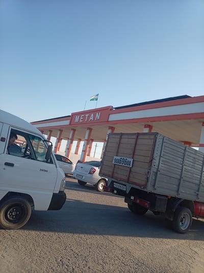 This media file depicts a street scene in Bagat, Uzbekistan, under a clear, bright blue sky, suggesting it is daytime. The central focus is a gas station with a prominent "METAN" sign displayed in large, red lettering on its facade. The gas station structure features a red and white color scheme, with supporting columns and a canopy. A flag, likely representing Uzbekistan, is visible flying from a pole above the gas station.
Several vehicles are present, indicating a possible queue or activity at the gas station. In the foreground, a white van, possibly a Damas Deluxe as indicated by text on its side, is parked, with its driver visible inside. To the right, a truck with a wooden cargo bed is positioned, with its license plate reading "90X696OA". Further back, a white car, identified by its license plate as possibly a Chevrolet, is also visible. The road surface appears to be asphalt with some granular texture. The overall atmosphere suggests a normal day at a local gas station, with everyday vehicles and infrastructure.