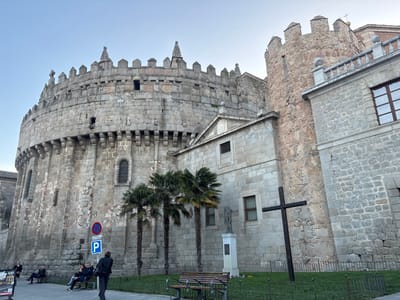 The image displays the fortified apse of the ÁVila Cathedral in ÁVila, Spain, integrated into the city walls, under a clear light blue sky. The prominent central and left portion of the scene is dominated by a massive, circular stone structure, the cathedral's apse, constructed from large, rough-hewn stone blocks. It features a crenelated parapet adorned with small, pointed spires and battlements, along with several narrow, arched windows.
To the right of the apse, adjacent stone buildings with varying textures of masonry are visible, including one section with a stone balustrade and windows, and another rougher stone tower-like structure extending upwards. Three tall, slender palm trees are positioned in front of the cathedral apse. Directly to their right, within a grassy area enclosed by a low metal fence, stands a light-colored stone statue of a figure on a pedestal. Further right, a large, dark wooden cross is planted upright in the grass.
In the foreground, a paved pedestrian area features several wooden benches. On the leftmost bench, three individuals are seated, and another person stands nearby. On a second bench to the right, two individuals are seated. A lone person in dark clothing walks away from the camera in the central foreground. A blue parking sign ('P') and a red circular 'no stopping' or 'no parking' sign are mounted on a pole near the palm trees. The overall environment suggests a public square or park area adjacent to the historic cathedral.