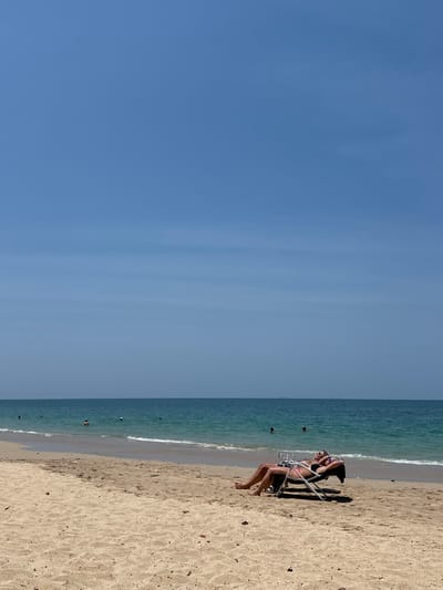 The image depicts a daytime scene on a sandy beach in Phra Ae, Thailand, under a clear, light-blue sky exhibiting faint horizontal cloud streaks. In the foreground, two individuals are positioned on separate, light-colored reclining beach chairs on the sand, oriented towards the sea. The individual on the right wears a dark two-piece swimsuit, sunglasses, and has arms placed behind her head. The individual on the left has visible tan limbs. The midground features the turquoise-blue sea with gentle waves breaking on the shore. Approximately six to eight dark figures, presumed to be people, are visible wading or swimming in the water, along with a few small white buoys. The sandy beach shows scattered minor debris and subtle impressions.