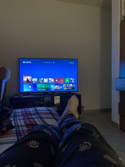 The image depicts an indoor scene in Arroyo Hondo, Mexico, captured from a first-person perspective, focusing on the lower body of an individual and a television display. The individual's legs are extended forward, resting on a plaid blanket featuring red, blue, and white patterns. They are dressed in dark blue pajama pants with a repetitive light-colored pattern and white socks.
Centered in the mid-ground is a large, black-framed flat-screen television, actively displaying the bright blue dashboard of an Xbox gaming console. The screen shows a user profile labeled "xavi varela oloya, xaviovg@gmail.com" and the time "07:44 p.m." Below the profile, various game and application icons are arranged, including "Fortnite" prominently displayed, "Juegos de simulación" (Simulation games), and "Suscríbete a Game Pass Ultimate" (Subscribe to Game Pass Ultimate). The television is positioned on a dark brown or black media console. Visible on the console are a white object resembling an Xbox console, a white collectible or game case featuring a "Halo" logo, and other indistinct items.
The room features light, neutral-colored walls. A portion of a light-colored furniture piece, possibly a headboard or armrest, is visible on the left. To the right, a section of dark wooden furniture with multiple drawers is present. The ambient lighting is dim, indicating an evening setting, with the television screen serving as the primary light source in the immediate viewing area.
