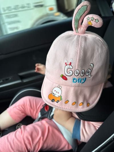 A baby is wearing a pink cap with bunny ears and embroidered decorations. The cap has stars, a carrot, a rabbit, and a small car with a bunny inside. The text "Good Day" is embroidered on the cap. The baby is wearing a pink long-sleeved shirt and pink pants. They are sitting in a car seat, possibly in a vehicle, as indicated by the dark interior and seatbelt. The baby's legs are bent. The image appears to be taken indoors, likely in a car during the daytime. The overall impression is one of a cute and comfortable baby.