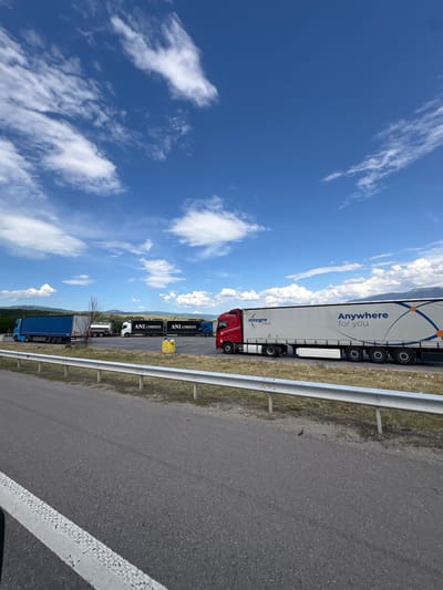 The image depicts a rest stop area along a highway, likely in the vicinity of Blatino, Bulgaria, under a bright blue sky with scattered white clouds. Several semi-trailer trucks are parked in the rest area. In the foreground, a gray asphalt road is visible, with a white painted line on the left edge. A silver metal guardrail runs along the edge of the road, separating it from the grassy embankment beyond. Behind the guardrail, several trucks are parked. On the left, a blue semi-trailer truck is parked. To its right, a white truck with a silver tanker trailer is parked. Further to the right, two black semi-trailer trucks with white lettering reading "ANI COMPANY" are parked side-by-side. A red semi-trailer truck with a white trailer is parked in the middle right of the image. The trailer of the red truck has the text "Integre trans" and "Anywhere for you" printed on it. In the background, hills and mountains are visible under the sky. A yellow cylindrical object is located on the ground near the parked trucks. The scene is static, with no visible people or activity.