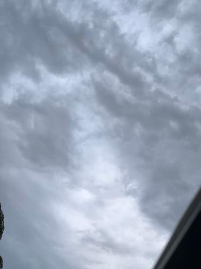 The image depicts a cloudy sky with overcast conditions. The clouds are predominantly gray, with some lighter patches indicating varying cloud density. In the lower left portion of the image, a dark, vertical object, possibly the foliage of a tree, is partially visible. The lower right corner of the image shows a dark, diagonal shape, likely the edge of a structure or rooftop. The scene appears to be an outdoor environment under a gloomy sky, with no people or specific activities visible. The location is identified as Kiyali-Kurgancha, Uzbekistan.