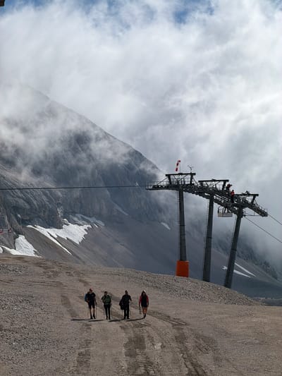 Four hikers are walking on a gravel path in a mountainous area with a ski lift in the background. The ski lift has multiple towers and cables. The mountain behind the ski lift is rocky and partially covered in snow, with clouds obscuring the top. The sky is overcast. One of the hikers is wearing a red jacket, another a green jacket, and the other two are wearing dark clothing. They are all using trekking poles. There are no visible indications that this scene is in Wildermieming, Austria.