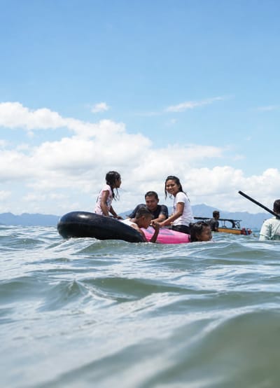 In the waters off Lumban Bul Bul, Indonesia, a group of people are enjoying a day on the water. A family is seated on inflatable tubes, with two children in the foreground and two adults behind them. The children appear to be playing, while the adults are smiling and looking at the camera. The water is calm and blue, with a few gentle waves. In the background, a traditional Indonesian boat is visible, along with lush green mountains. The sky is a bright blue, with a few fluffy white clouds. The overall scene is one of joy and relaxation, with the family enjoying their time together in the beautiful Indonesian landscape.