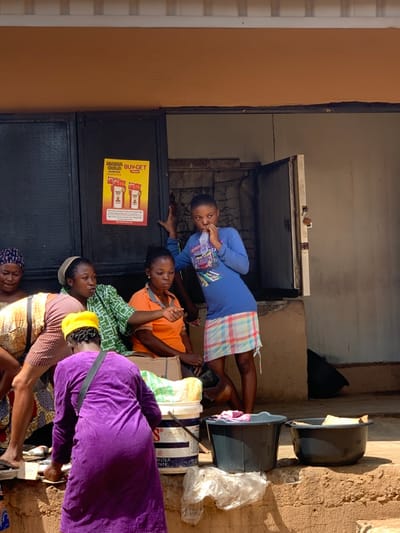 The image depicts a scene in front of a building in Ondo, Nigeria. The coordinates 7.251, 5.196 place the location in the city of Ondo, close to the main market. Four women are interacting in front of the building. One of the women is wearing a purple dress and is seated on the ground, possibly selling something. She is looking at the ground. Another woman is wearing a yellow dress and is seated on a concrete step. She is talking to a woman wearing a green and white dress. The fourth woman is standing and is looking towards the right. She is wearing a blue shirt and a plaid skirt and is drinking from a bottle. The women are all African in appearance. There is a poster on the wall behind them that says "Burger Gold Buy Get". The environment is a typical urban scene with a building, a concrete step, and a few trash cans.