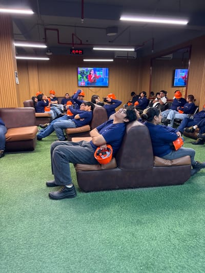 The image depicts a group of people, most of whom are wearing dark blue shirts and orange safety helmets, seated on brown leather couches arranged in a semi-circle. The environment appears to be an indoor waiting area or lounge with a green carpet resembling artificial grass. In the background, there are several screens displaying content, and a digital clock on the wall shows "10:26:24". The people are casually seated, some with their eyes closed, suggesting they might be resting or waiting. One person in the foreground is holding an orange helmet. There is no indication of the city of Villa El Salvador, Peru within the image itself.