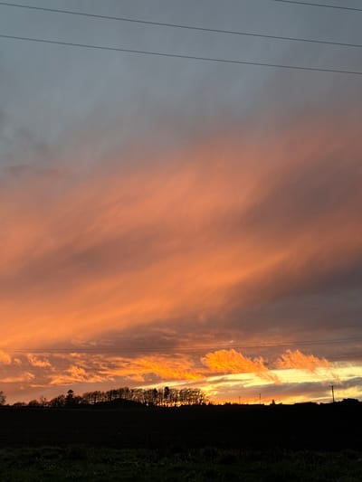 The image shows a landscape in Fowlis Easter, United Kingdom, at dusk or dawn. The sky is predominantly orange with cloud formations. To the left, there are greyish-blue skies. Power lines are visible against the sky. On the right, there is a silhouette of a treeline and green vegetation.