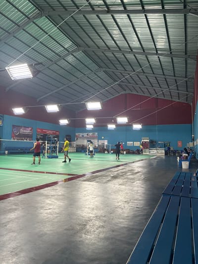 This indoor sports facility, located in Kecamatan Cisaat, Indonesia, showcases an active badminton hall. The scene is dominated by multiple green badminton courts, clearly marked with white boundary lines and bordered by maroon-colored strips.
Several individuals are present, engaging in or observing badminton. In the foreground, two male players, one in a red shirt and the other in a yellow shirt, are positioned on a court, holding rackets and appearing ready to play. Further back, more players are visible on an adjacent court. Along the far wall, additional people are seated on blue benches, possibly spectators or waiting for their turn.
The setting is a large hall with a high, arched, corrugated metal roof, supported by visible metal beams, giving it an industrial aesthetic. Numerous bright, rectangular fluorescent light fixtures are suspended from the ceiling, providing strong illumination to the courts. The walls are painted in segments of blue and red. Along the right side of the image, several rows of empty blue bleachers indicate spectator seating.
The activity clearly revolves around badminton, with players actively participating or preparing to play. The artificial lighting suggests the time of day is either evening or night, or simply that the indoor nature of the facility makes natural light less relevant.
Visible text on a banner along the back wall includes "AMKA", "Jaya", and what appears to be "Salam Olahraga!", along with other smaller, less discernible text, likely pertaining to a club, event, or sponsor. The overall impression is one of an energetic community sports center.