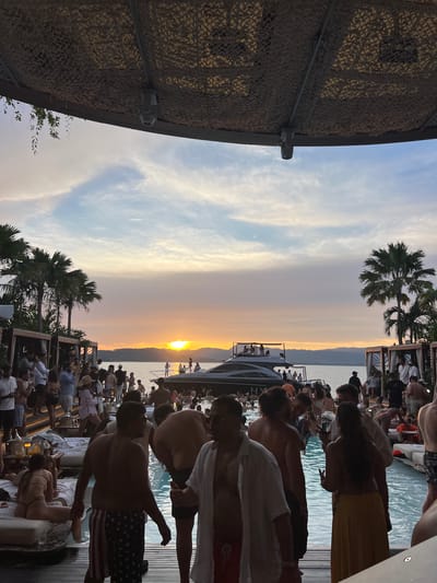The image depicts a lively outdoor party scene at a resort in Pakhlok, Thailand, during sunset. A large group of people are gathered around a swimming pool and on a wooden deck, with the ocean and distant hills visible in the background. Several boats are moored in the water, and a yacht is prominently positioned near the shore. Palm trees line the edge of the property, adding to the tropical ambiance. Attendees are dressed in casual, summery attire, with many in swimwear. The atmosphere appears festive and relaxed, with people socializing and enjoying the evening. The setting sun casts a warm, golden glow over the landscape, creating a beautiful and serene backdrop for the celebration. The resort features cabanas and lounge areas, indicating a luxurious and well-equipped venue.