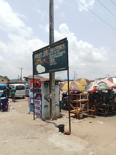The image shows a street scene in Ovwian, Nigeria. A large green sign advertising "Sunny Eru Motors" is prominently displayed on a metal pole, listing destinations including Bayelsa, Yenagoa, Port Harcourt, Aba, Agbor, and Onitsha. The sign also includes the words "Udu Route" and phone numbers. The scene is dusty and sandy, with a few cars parked nearby. Several people are present, including a few standing near the parked cars and others seated at tables under umbrellas. To the right of the sign is a small, partially visible structure with a sign that says "Available Here." This structure is decorated with advertisements for bank services including "deposit," "withdrawal," and "transfer."