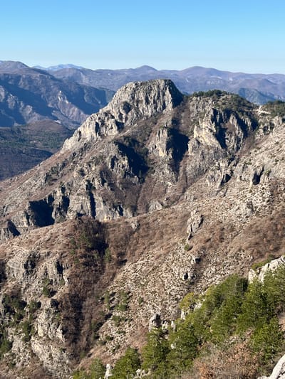 The image depicts a vast, rugged mountainous landscape under a clear, pale blue sky, located in the vicinity of Klobuk, Bosnia and Herzegovina. The foreground and midground feature steep, rocky slopes characterized by exposed gray and brown rock formations, interspersed with sparse vegetation. Patches of evergreen trees, primarily pine, appear in the lower right and along some ridges, contrasting with areas of deciduous growth that are largely bare or possess minimal brown foliage. A prominent, jagged rock peak dominates the mid-left, exhibiting significant erosion and vertical fissures. Multiple layers of mountains extend into the background, becoming progressively hazier and bluer, indicating considerable distance. Sunlight illuminates the scene, casting shadows that accentuate the three-dimensional topography of the terrain. No human activity, structures, or animals are visible.