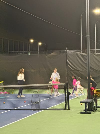 A sideways view of a tennis court at night. Court lights illuminate the scene. Multiple people are on the court, including adults and children. One adult appears to be coaching a child. Tennis balls are scattered around the court. A net divides the court, and a ball collection cart is visible. Fencing surrounds the court.