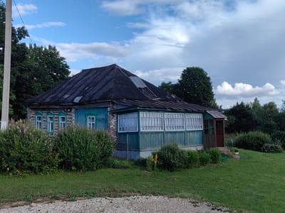 The image captures a weathered, single-story house with a dark, corrugated metal roof in the SNT Romashka, Russia. The house has light blue walls and a green, enclosed porch with multiple windows that are covered with white curtains. To the left of the house, a dense bush with pink flowers borders the property, and to the right, a grassy lawn slopes gently downwards. A gravel path runs along the bottom of the image. The sky is partly cloudy with a mix of blue sky and grey clouds. Trees surround the property, creating a rural or semi-rural setting. There are no people or animals visible in the frame.