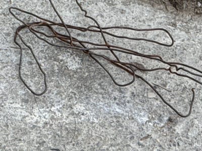 This is a close-up, abstract shot of tangled, dark, thin wires against a textured, light gray, granular background. The wires are bent and twisted into various shapes, forming loops, curves, and straight segments. Some of the wires appear to be intertwined with each other, creating a complex network. The background resembles a rough concrete or stone surface, with a speckled appearance due to its granular texture.
The lighting appears to be diffuse, with no harsh shadows, suggesting an overcast day or indirect light. The overall mood is somewhat industrial or utilitarian, focusing on the form and texture of the materials. There is no visible text in the image, nor are there any people or overt signs of activity. The image is purely an exploration of line, form, and texture, with the wires being the primary subjects against the contrasting background. The location context of Mastaga, Azerbaijan, does not offer specific visual cues within the image itself that would tie it to a particular landmark or characteristic of that region, beyond the general impression of an outdoor or construction-related environment.