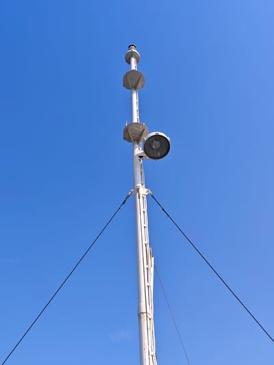 A tall, white mast extends upwards against a clear blue sky. The mast is equipped with several features, including what appears to be a light fixture and navigational signals. Guy wires are attached to the mast, anchoring it to unseen points below. The overall scene is devoid of any people or additional objects, suggesting a focus on the solitary structure against the vast expanse of the sky. The location is identified as Novonazimovo, Russia.