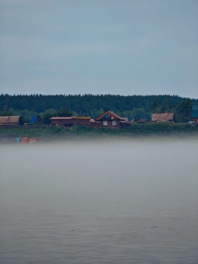 The image depicts a foggy morning scene along the banks of a body of water, likely a river, in the village of Atamanovo, Russia. In the foreground, the water's surface is visible, characterized by gentle ripples and a muted gray color. A thick layer of fog obscures the lower portion of the scene, creating a hazy transition between the water and the land.
Beyond the fog, on the elevated bank, a collection of rustic wooden houses and other structures are visible. These buildings appear to be older, with weathered wooden facades and a mix of pitched and flat roofs. Some of the houses have windows, and one house on the left has a distinct red-tiled roof. There are also what appear to be storage containers or small structures in a rust color located near the water's edge, partially shrouded by vegetation.
Behind the village, a dense forest of dark green coniferous trees forms a backdrop, stretching across the horizon. The sky above is overcast and a pale gray, contributing to the subdued and slightly somber atmosphere of the scene. The overall impression is one of a quiet, rural settlement on a misty morning.