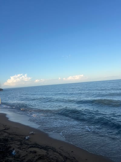 The image depicts a coastal scene. A clear blue sky occupies the left portion, transitioning into a lighter hue towards the horizon. Fluffy white clouds are visible along the horizon line, separating the sky from the ocean. The ocean extends towards the right, with visibly choppy water. The water meets a sandy beach on the right side of the image, where some organic debris are scattered. The image is rotated counterclockwise. It is impossible to confirm the location in Dowh, Malawi based on the provided image. No people, subjects, or interactions are directly observable in the scene.