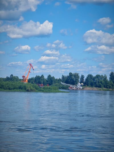 A wide, calm river flows in the foreground of an image taken on a sunny day in Krutoi Log, Russia. The sky is a vibrant blue, dotted with fluffy white clouds. On the far bank, a dense line of lush green trees forms the backdrop. To the left, a tall, orange construction crane stands prominently, its boom extended. Further to the right, a white barge with a crane is moored on the water near the shore. The scene appears to be a quiet industrial or port area, with the natural landscape dominating the view. There are no people visible in the frame.