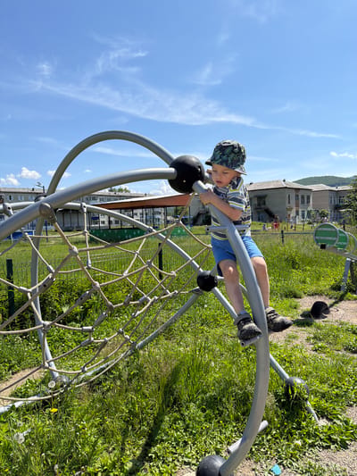In the city of Kuruldur, Russia, on a bright, sunny day, a young boy is playing on a jungle gym. He is wearing a bucket hat, a striped t-shirt, and blue shorts, with sturdy shoes on his feet. The jungle gym is made of metal bars and ropes, forming a large, spherical net. The boy is in the process of climbing, holding onto a metal bar and resting his feet on another. The surrounding area is a playground, with more play equipment visible in the background, along with residential buildings and a hill. The ground is covered in green grass and dirt paths, with dandelions and other wildflowers scattered about. The sky is a clear blue with a few wispy clouds.