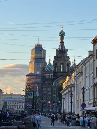 The image captures a street scene in the city of St. Petersburg, Russia, featuring the iconic Church of the Savior on Spilled Blood. The church's distinctive onion domes are visible, one of which is adorned with green and blue stripes. A tall, octagonal tower, partially covered in scaffolding and reflective material, stands adjacent to the church. The sun, low in the sky, casts a warm golden light on the buildings and clouds.

The street is lined with classical European-style buildings with ornate facades. A sidewalk bustles with pedestrians, some walking, some gathered in small groups. Several street lamps, tall and black, illuminate the path. Traffic signs are visible, including a pedestrian crossing sign and a "no stopping" sign. A stone bridge or embankment is in the foreground, with people standing along its edge. The sky is a clear blue, with a few wispy clouds present. The overall impression is of a vibrant, historic urban setting bathed in the soft light of late afternoon or early evening.
