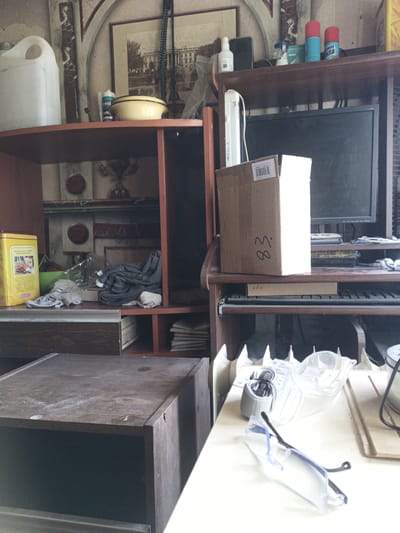 The image depicts an indoor scene, likely a room in a residential or workshop setting in Dobryatino, Russia. The room is cluttered with various objects on shelves and surfaces, suggesting a state of disarray or storage. Prominently featured are shelves containing items such as a large white plastic container, a yellow tin, grey cloth items, a computer monitor, a cardboard box with markings, and a keyboard. The overall environment appears to be dusty and unorganized. There are no people visible in the image, and no specific actions or interactions are evident. The background shows decorative architectural elements, possibly wallpaper or painted designs, contributing to the overall aesthetic of the space.