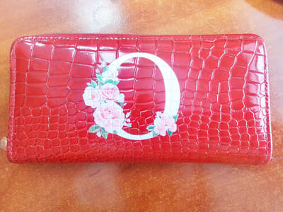 The image shows a red wallet with a crocodile-like texture. A white letter "O" is printed on the front of the wallet, partially covered by a floral design featuring pink and white roses with green leaves. The wallet is lying on a wooden surface, likely a table. The background is blurred, suggesting a shallow depth of field, and the lighting is bright, highlighting the glossy texture of the wallet. There are no people or animals present in the image. The image does not provide any specific information or context related to the city of Сарыкөл, Kazakhstan.