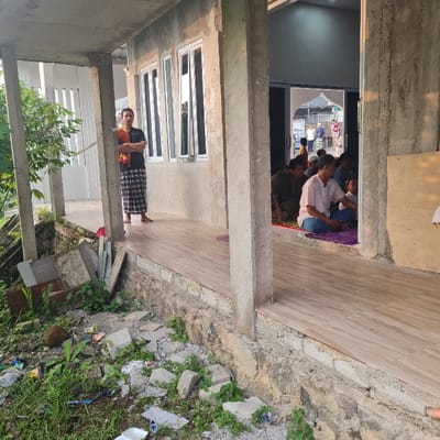 The scene is an outdoor patio of a building in Kecamatan Tamansari, Indonesia, at latitude -6.635 and longitude 106.738. The patio has a tiled floor, a wall on the right side and a partially covered walkway to the left. A man in a dark blue shirt and brown pants stands on the tiled floor, facing left. In the background, there are several people seated in a room with a glass door. The people are talking and looking at each other. The room is lit by overhead lights. The room has windows on either side of the glass door. There are trees and other vegetation outside the patio. Rubble and debris are scattered on the ground around the patio. The scene is daytime.