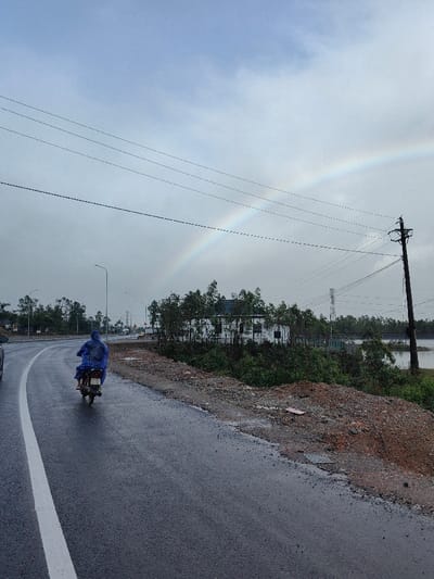 The image depicts a rural or semi-urban roadway scene in Hoành Sơn Ward, Vietnam, under an overcast sky with a prominent rainbow. A single individual, clad in a blue rain poncho, operates a motor scooter away from the viewer on the wet asphalt road, which curves slightly to the left. A solid white line marks the left edge of the visible road. To the right of the road, an unpaved shoulder of reddish-brown dirt and gravel slopes up to an area dense with green foliage and trees, partially obscuring a white building. A utility pole with multiple power lines stands on the right, connecting to lines that stretch across the upper portion of the frame, passing through the visible rainbow arc. In the background on the right, beyond the trees, a body of water is visible. On the left, a portion of another vehicle (car) is partially visible, and further down the road, streetlights and additional trees line the route.