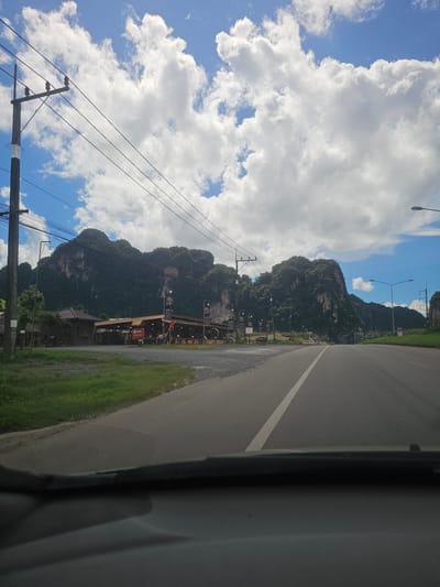 The media file depicts a scenic view along a road in Ao Luek, Thailand. The primary subjects are the dramatic, lush green limestone mountains that dominate the background, interspersed with patches of blue sky and fluffy white clouds. A paved road, marked with a white line, runs alongside a grassy verge. Overhead power lines and utility poles are a prominent feature in the foreground and mid-ground, crossing the sky and extending towards the mountains.

Several structures are visible nestled at the base of the mountains, suggesting a small community or a roadside commercial area. These include buildings with reddish roofs and some open-air establishments with tables and chairs. A few lampposts are strategically placed along the roadside. The overall impression is of a rural or semi-rural setting in a tropical climate, likely during the daytime given the bright sunlight and clear blue sky.

There is some text visible on signage, but it is not clearly legible. The weather appears pleasant, with a mix of sun and clouds. The perspective suggests the image was taken from inside a vehicle, looking out towards the landscape. The scene evokes a sense of journey and exploration through a beautiful natural environment.