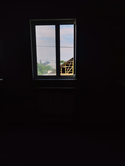 A rectangular, white-framed window is centrally located in a dark, interior room. The window is divided into two panes. Through the left pane, a cloudy sky and green trees are visible, along with power lines stretched horizontally across the frame. The right pane offers a view of a partially constructed building with a wooden frame, its roofline visible. Below the window, a dark-colored radiator with horizontal fins is positioned. The walls of the room are a dark, muted color, making the window the focal point of the image. The scene suggests an indoor perspective looking out onto a residential or semi-rural area. The location of Zhanadaur, Kazakhstan is not depicted or indicated in the image.