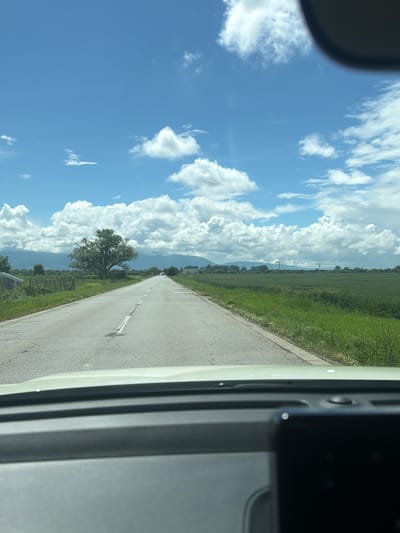 The image, taken from inside a vehicle, captures a scenic view of a road stretching into the distance under a bright, cloud-filled sky. The perspective is looking forward and slightly to the left, with parts of the vehicle visible on both sides of the frame.

In the foreground, a paved road with a white broken center line and a solid white edge line extends towards the horizon. To the right of the road, green grass and fields are abundant, with a small patch of possibly wild red flowers visible. To the left, similar green fields line the road, featuring a large, solitary deciduous tree with a full canopy.

The mid-ground reveals more expansive green fields, suggesting an agricultural or rural landscape. In the background, distant mountains or hills rise gently, partially obscured by the clouds or atmospheric haze. The sky is a vibrant blue, filled with large, fluffy cumulus clouds that stretch across the horizon, indicating fair weather.

The time of day appears to be late morning or afternoon, with ample sunlight illuminating the scene. The overall impression is one of a peaceful drive through a rural countryside. No visible text is present in the image.