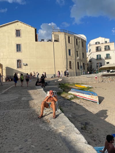 This image depicts a sunlit, bustling scene in the coastal town of Cefalù, Sicily, Italy.
In the immediate foreground, to the left, a bald, tanned male, appearing middle-aged to elderly, sits on a white, collapsible beach chair. He wears dark sunglasses, patterned blue, yellow, and white swim trunks, and black flip-flops. His skin has a sheen, suggesting moisture or lotion. He wears a watch on his left wrist and a blue bracelet on his right. He is positioned on a sloped, cobblestone path or plaza, which constitutes the lower-left quadrant of the frame. To his immediate right, a concrete ledge or retaining wall separates the cobblestone area from a sandy beach.
The midground features this sandy beach, where two boats are visible. One, larger, is blue with faded red and white accents, lying overturned on the sand. A smaller, yellow kayak or canoe rests upright nearby. Further to the right, a person is partially visible sitting on a blue towel on the sand, facing away from the camera. Shadows from unseen objects or structures are cast across the sand.
Behind the man in the chair and extending across the mid-ground, a series of stone or concrete steps ascends from the cobblestone area towards a higher level. Approximately 15-20 individuals are dispersed across these steps and the upper cobblestone path, walking, standing, or interacting in small groups. Their attire is casual summer wear. A small child in a red top is visible on the beach steps in the mid-right.
The background is dominated by multi-story buildings characteristic of Mediterranean architecture. The closest building on the left features a light cream-colored facade with several rectangular windows, some with visible vertical cracking on the wall surface. An air conditioning unit is affixed to its lower section. A rooftop terrace with metal railings and outdoor seating is visible atop a connecting structure. Further right, another light-colored building with balconies and green-shuttered windows completes the urban backdrop. The scene is illuminated by strong sunlight, casting defined shadows. The sky above is clear blue with scattered white clouds.