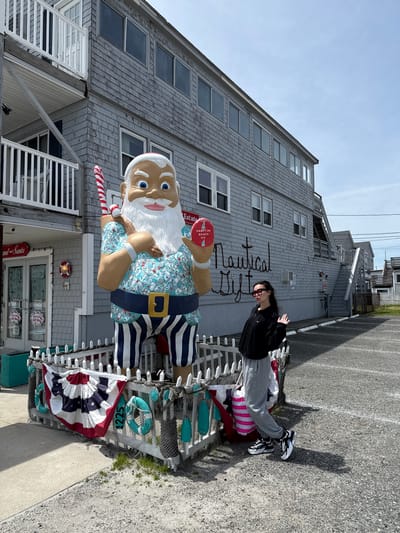 This outdoor image, taken in Hampton, United States of America, depicts a large, colorful, nautical-themed Santa Claus statue positioned in front of a grey shingled building. A person is posing casually next to the statue.
The central subject is a tall, cartoonish Santa figure with a white beard and blue eyes, dressed in a light blue patterned shirt, striped blue and white pants, and a dark belt with a gold buckle. He holds a red and white candy cane in his left hand and a red object with "Hampton Beach N.H." written on it in his right. The statue is situated behind a white picket fence adorned with fishing nets, blue and white buoys, and life rings, one of which prominently displays "1225". A red, white, and blue patriotic bunting is draped over the fence towards the bottom right.
To the right of the statue, a person with dark hair, wearing sunglasses, a black jacket, grey sweatpants, and black and white sneakers, stands with their hands raised near their face, looking towards the left.
The background features a multi-story building with light grey shingle siding and a steep roof, characteristic of coastal architecture. The words "Nautical Gifts" are prominently displayed in white script on the building's facade. Below the Santa statue, a storefront entrance with an "OPEN" sign and "Gift Shop" text is visible. The ground is asphalt pavement, suggesting a parking area or sidewalk. The scene is illuminated by bright daylight under a clear, light blue sky, with visible shadows indicating the sun's position. The overall impression is of a vibrant, themed gift shop or attraction at a seaside location.