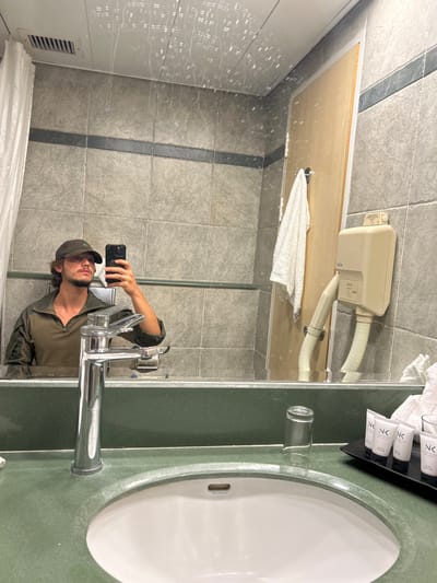 This image depicts a mirror selfie taken by a male in a hotel bathroom, located in Tiberias, Israel.
The male subject, reflected in the mirror, possesses medium-length dark hair, a beard, and is attired in a dark green baseball cap and a dark olive-green long-sleeve shirt with a zippered collar. He is holding a black smartphone with a visible camera array in his right hand, positioned to capture the reflection.
The bathroom environment includes a dark green countertop featuring a white oval-shaped sink and a chrome single-lever faucet. To the right of the sink, an inverted clear glass cup is present, alongside a black rectangular tray holding four small white tubes of toiletries labeled "NK". The walls are tiled with light grey square tiles, visually divided by a horizontal dark grey border stripe. Above the reflection of the subject, a white ceiling with a rectangular ventilation grate is visible. The mirror surface exhibits visible streaks consistent with water droplets or condensation. Further elements reflected include a light-colored wooden door with a white towel hanging from a hook, a beige wall-mounted hair dryer unit with a flexible hose, and a partial white shower curtain on the far left.