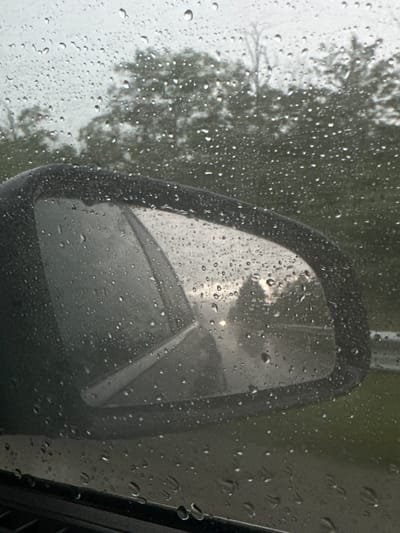 The image captures a close-up view from inside a vehicle, looking out through a window heavily adorned with raindrops. The primary subject is the vehicle's black-framed side mirror, positioned towards the center-right of the frame, also covered in water droplets.

Through the rain-splashed window, a blurred, soft-focused background of green foliage, likely trees or bushes, is visible, suggesting an outdoor, natural setting. The individual raindrops on the window vary in size and shape, creating a textured, diffusing effect over the view beyond.

The side mirror reflects a somewhat indistinct scene, characterized by muted grey and dark tones, with a notable bright, almost glowing, spot towards the upper part of its reflection. This bright area could indicate the sun attempting to break through clouds or another bright environmental element. A dark, angular part of the car's frame or interior is visible along the right edge of the image.

The scene depicts a rainy or recently rainy day, observed from the shelter of a vehicle. The lighting suggests it is daytime, possibly overcast. No people or visible text are present. The setting is Zvezditsa, Bulgaria.