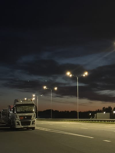 A white semi-truck is parked on the right side of a highway at dusk or night. The sky is dark with some clouds. There are several streetlights visible along the side of the highway. The truck appears to be stationary and its headlights are on. The road is paved with white lane markings. The background shows a line of trees and what appears to be a building or structure. The overall scene is dimly lit, suggesting it is either late evening or early morning. The image is oriented vertically.