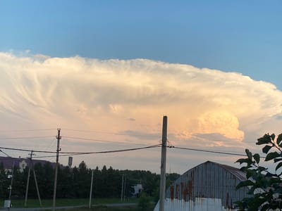 The image shows a large, anvil-shaped cloud formation in the sky over a rural landscape. The cloud is illuminated by the setting sun, casting a warm, golden glow on its underside. The sky is a clear blue, transitioning to a pale pink and orange near the horizon.
In the foreground, there are several power lines supported by wooden and concrete poles. Behind the power lines, a line of dark green trees stretches across the middle ground. To the left of the image, a building with a purple roof is visible, and further to the right, a dilapidated, corrugated metal structure with rusted panels is partially visible. The ground in front of the trees appears to be a grassy field.
The scene is located in the village of Beskol in the Beskol Rural District of Kazakhstan. The image captures a moment of natural beauty, with the dramatic cloud formation dominating the sky and contrasting with the mundane elements of the rural infrastructure. There are no people or animals visible in the image. The overall impression is one of quiet, expansive natural beauty tinged with the signs of human presence in the form of utility infrastructure and aging buildings.