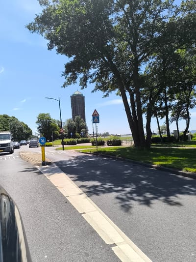This is a street-level, eye-level, medium shot, outdoor photograph taken on a sunny day in Aalsmeer, Netherlands. The image features a road with a painted yellow line marking the edge and a white crosswalk. In the background, there's a prominent, multi-story, dark-colored water tower with a green dome, surrounded by trees and lush green grass. Several cars are visible on the road, and there are traffic signs present, including a pedestrian crossing sign. The scene is characterized by clear blue skies and shadows cast by trees on the road.